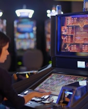 A person is seated at a slot machine console in a casino, focusing intently on the game displayed on the screen. The background shows several other illuminated gaming machines, adding a vibrant and colorful atmosphere to the scene.